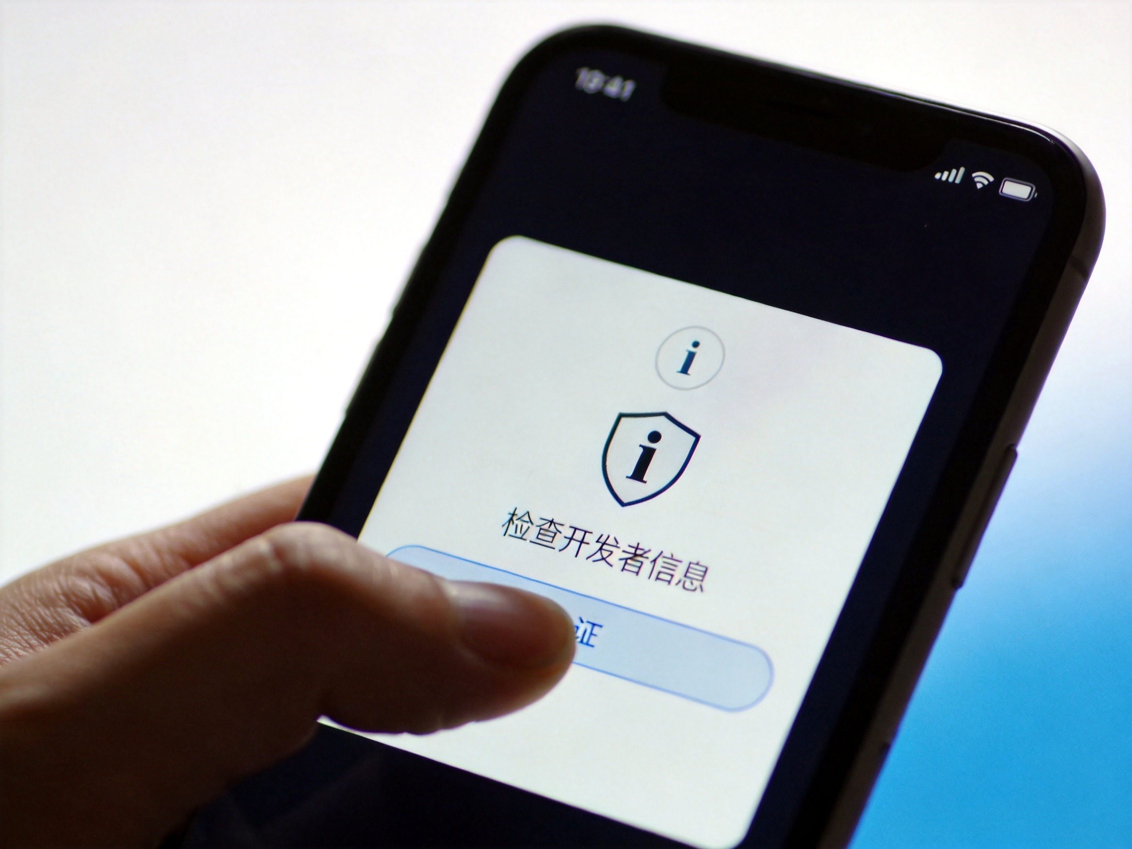 GEO正文图二：手机屏幕特写，指尖点击屏幕上的信息验证区域和盾牌认证徽章，强调核对开发者身份。